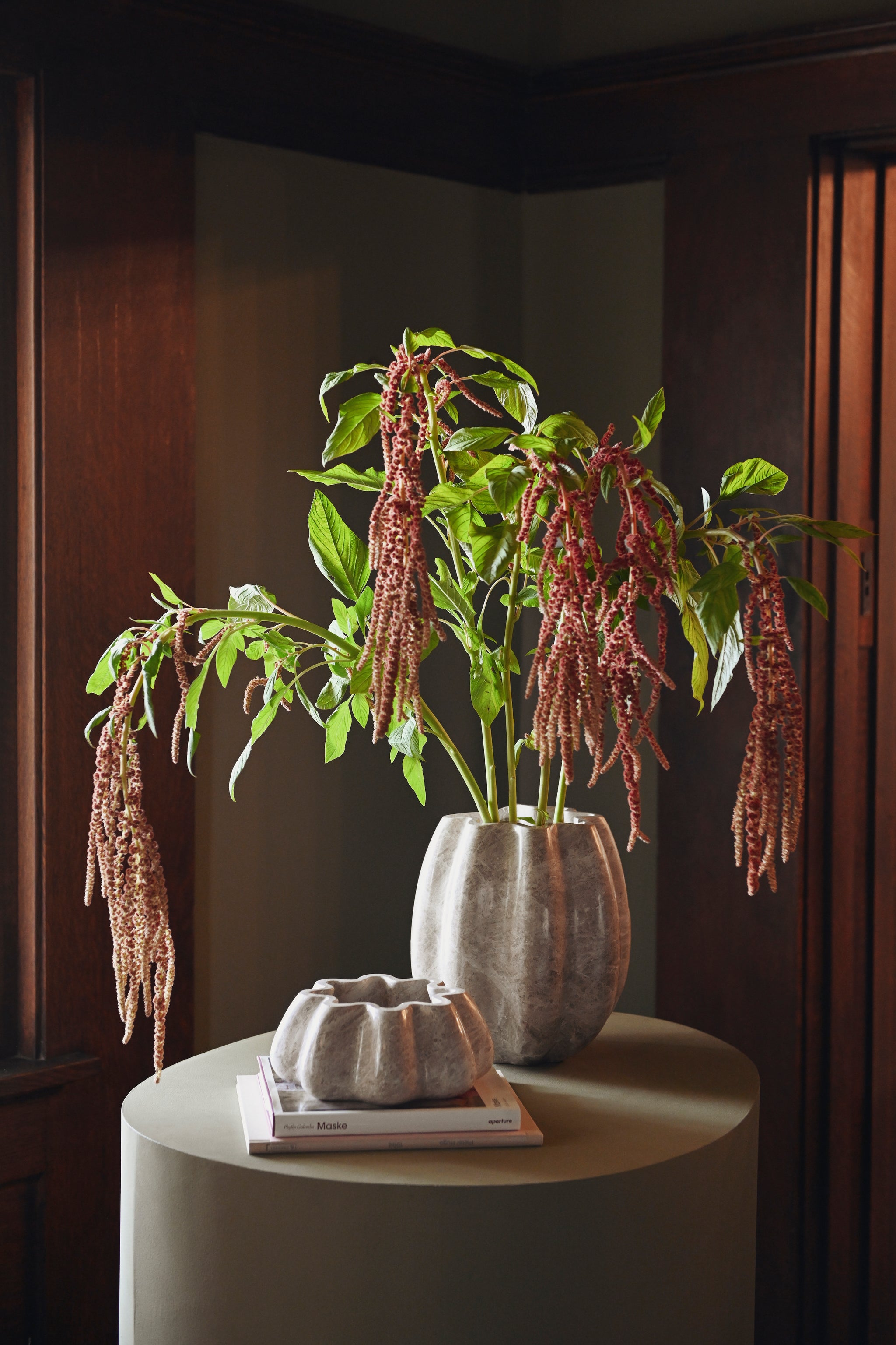 A tall oyster marble vase of trailing amaranth and a fluted oyster marble bowl on a pedestal table in a paneled room