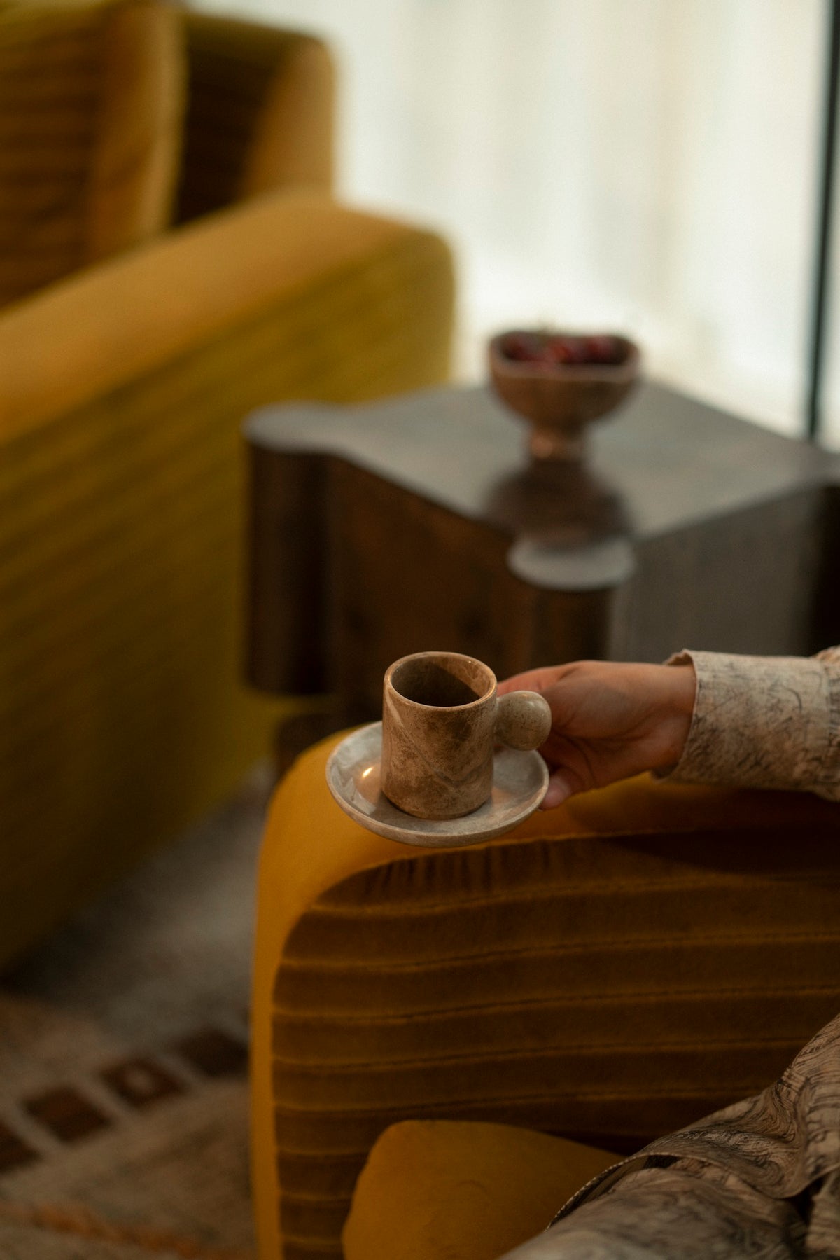 A hand setting a marble cup on a side table beside a citron armchair