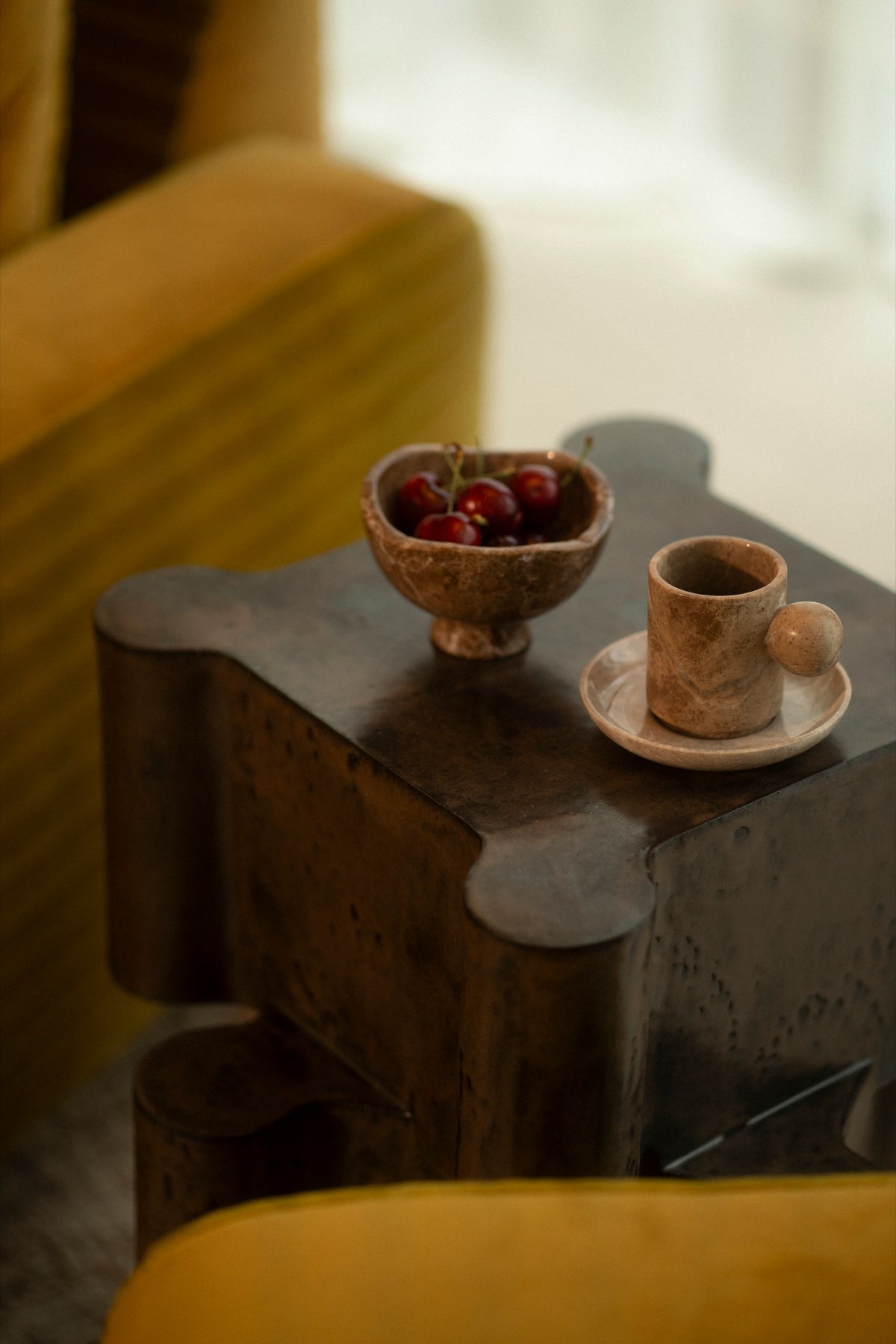 The Date Bowl filled with cherries and a marble cup on a dark side table between citron armchairs