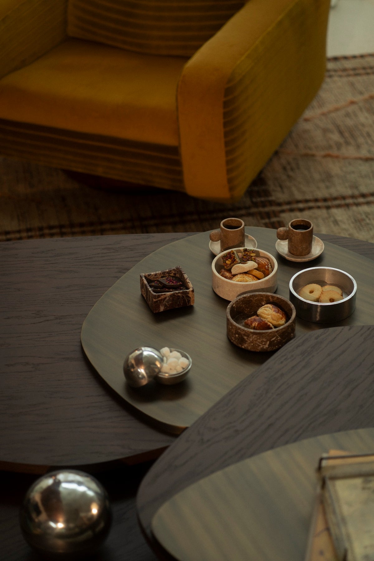 Marble bowls of pastries and cups on a dark table below a citron armchair