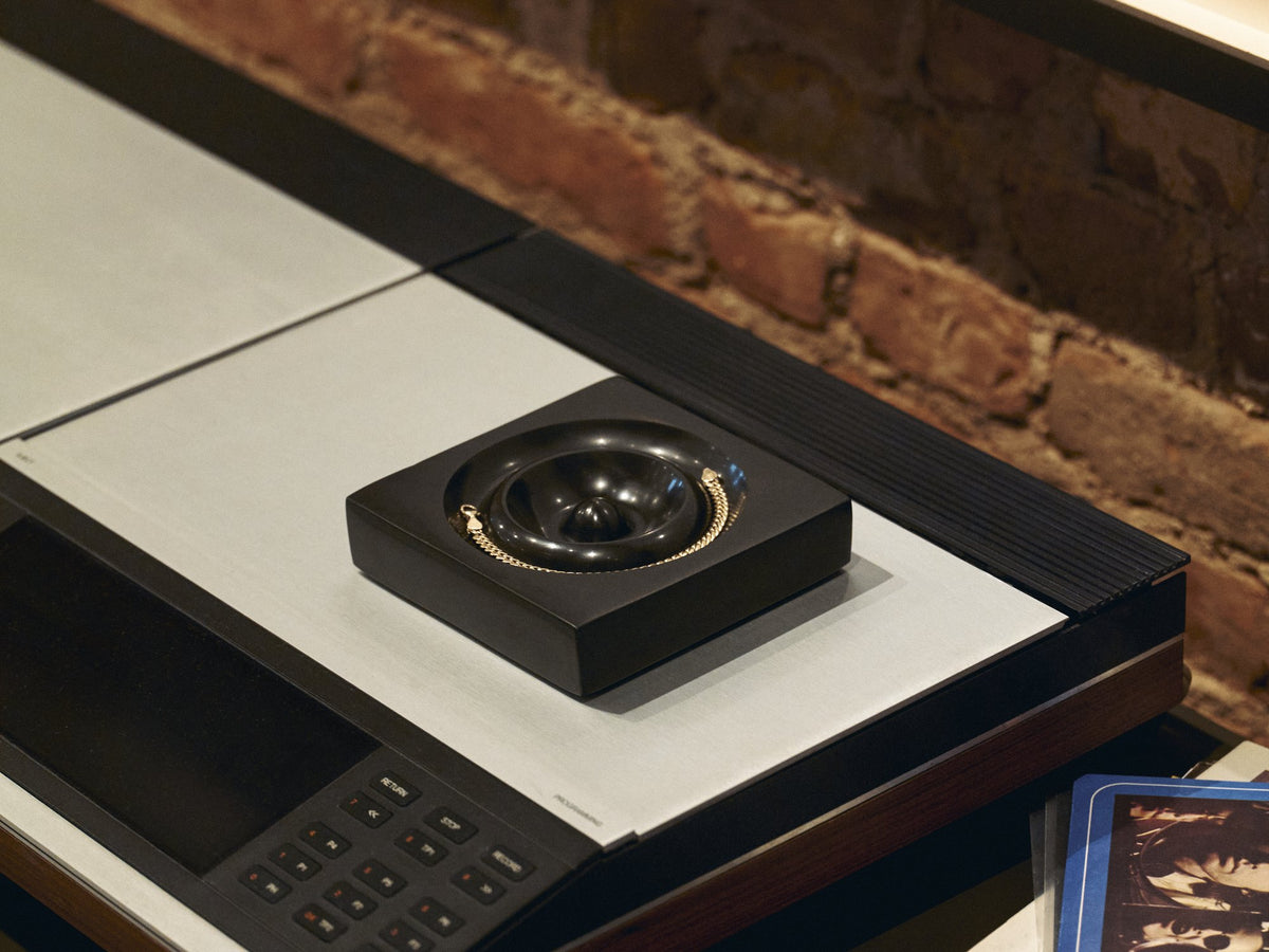 A black marble catchall holding a gold chain on a hi-fi console against a brick wall