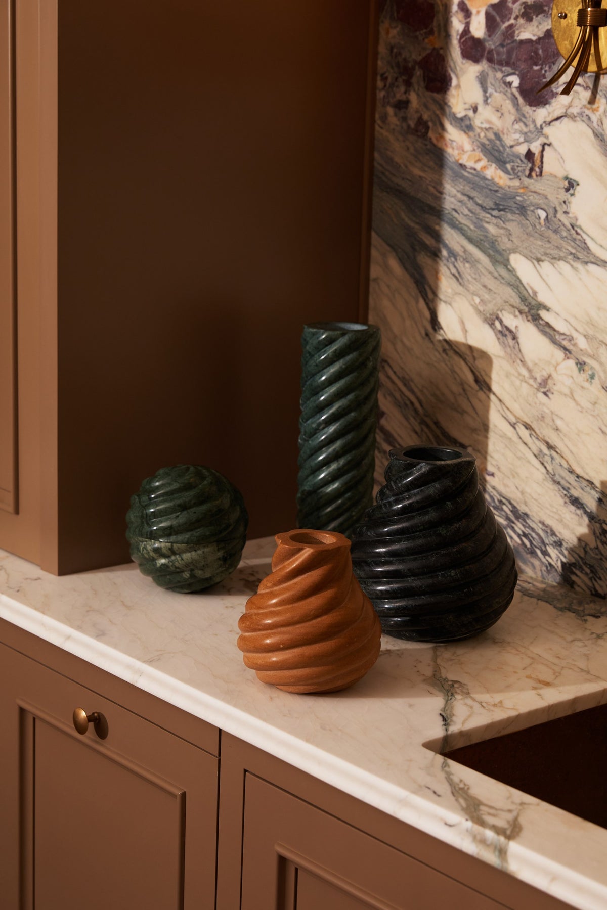 Four Swell vases together on a marble kitchen counter: a round and a tall twisted vase in Emerald, a large vase in black, and a small vase in terracotta stone